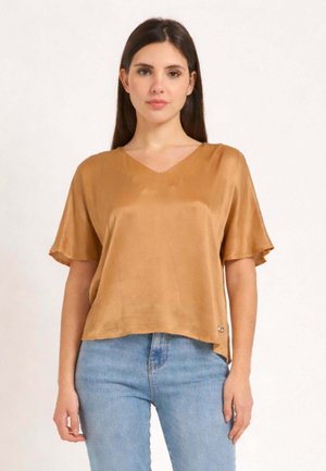 Donna che indossa una camicetta dorata in seta, ampia, a maniche corte e scollo a V, abbinata a jeans di denim azzurro chiaro, in piedi su uno sfondo semplice.