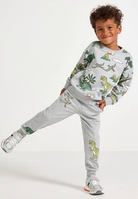 Smiling boy stands on one leg wearing grey sweatshirt and pants with green dinosaur and volcano prints and white sneakers.