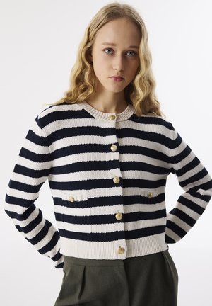 Marineblauer und cremefarbener gestreifter Cardigan mit Rundhalsausschnitt, goldenen Knöpfen und Fronttaschen, kombiniert mit olivgrünen Hosen. Weiche Stricktextur.