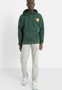 Donkergroene hoodie met een voorkantzak en een hartgrafiek, gecombineerd met lichtgrijze cargobroek met meerdere zakken en sneakers.