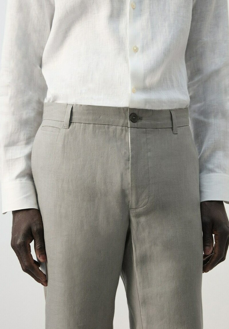 Homme portant un pantalon gris clair et une chemise blanche à manches longues boutonnée, debout avec les mains détendues le long du corps.