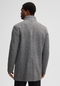 Cappotto in lana grigia a spina di pesce con colletto alto, spalle strutturate e vestibilità dritta, caratterizzato da cuciture sul retro e dettagli hardware minimi.