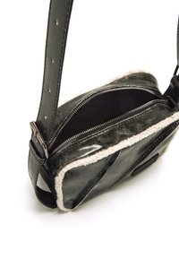 Bolso cruzado de cuero negro con una superficie texturizada, ribete de borreguito blanco, cierre superior con cremallera y una correa ajustable con herrajes metálicos.