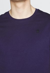 Ylävartalo henkilöstä, joka käyttää pelkkää tummanliilaa pyöreäkaulaista t-paitaa, jossa on pieni musta logo vasemmalla rinnassa.