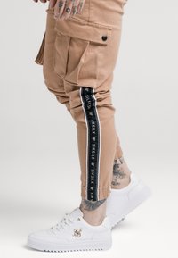 Beige cargobroek met elastische manchetten, voorzien van zwarte zijstrepen en logodetail. Draagt witte sneakers die een textuur oppervlak tonen.