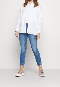 Vit skjorta med knappar, matchad med ljusblå tapered jeans med cargo-fickor och elastiska ärmslut, kompletterad med beige sneakers.