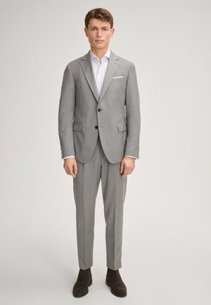 Młody mężczyzna stoi, ubrany w jasnoszary garnitur na dwa guziki, białą pasiastą koszulę i ciemnobrązowe buty na jednolitym jasnym tle.