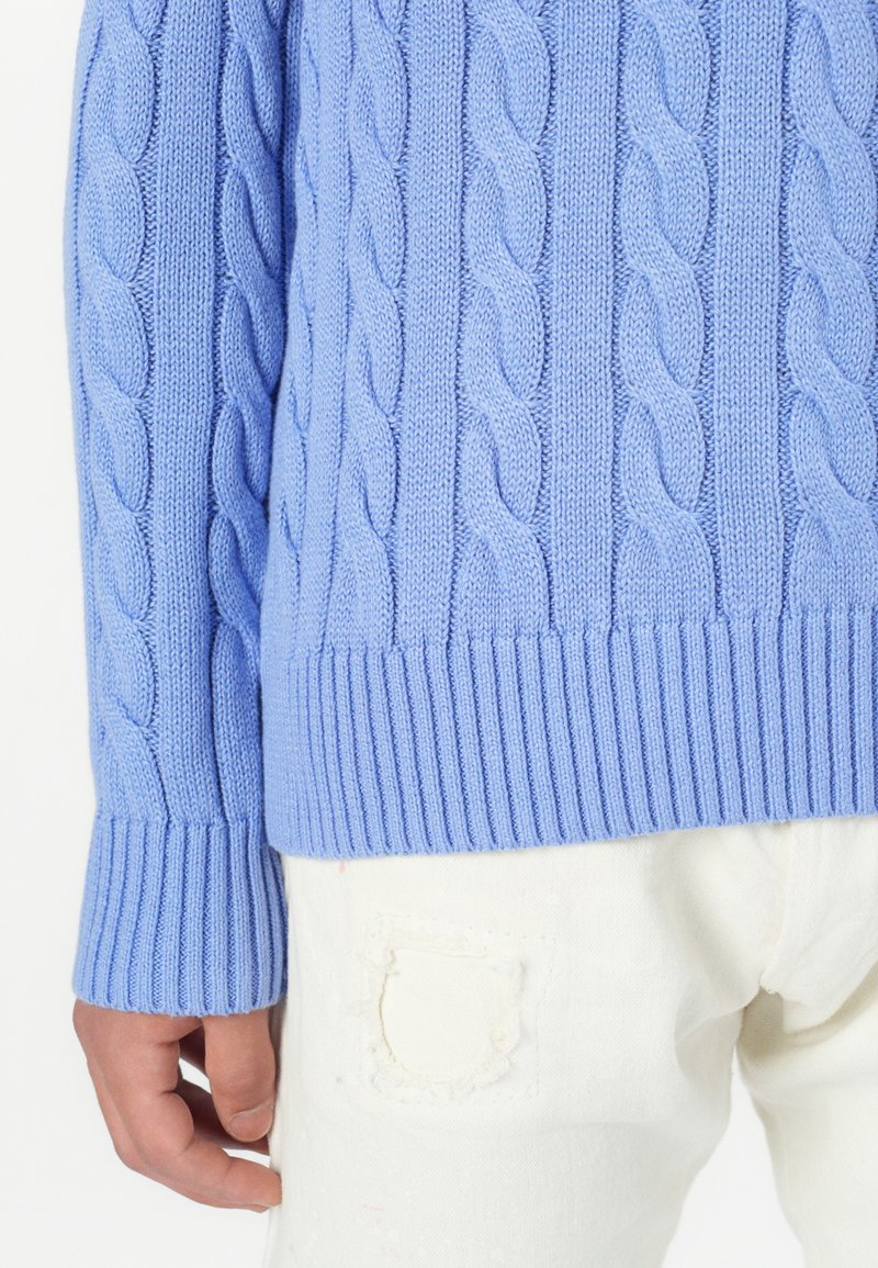 Maglione azzurro chiaro a maglia rialzata con polsini e orlo a coste, abbinato a pantaloni bianchi con una toppa strappata al ginocchio.