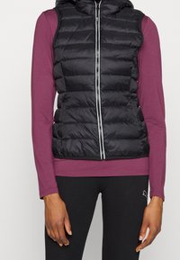 Svart pufferväst med huva, med horisontellt quiltat mönster, framtill dragkedja, bärs över en långärmad burgundskjorta och svarta leggings.