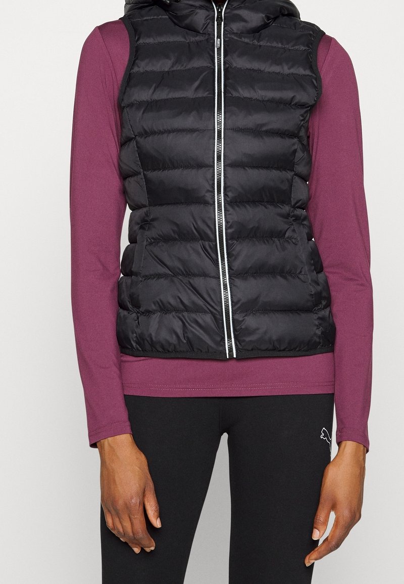 Svart pufferväst med huva, med horisontellt quiltat mönster, framtill dragkedja, bärs över en långärmad burgundskjorta och svarta leggings.