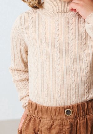 Enfant portant un pull en maille torsadée crème à col roulé et un pantalon en velours côtelé marron avec une ceinture boutonnée, main près du visage.