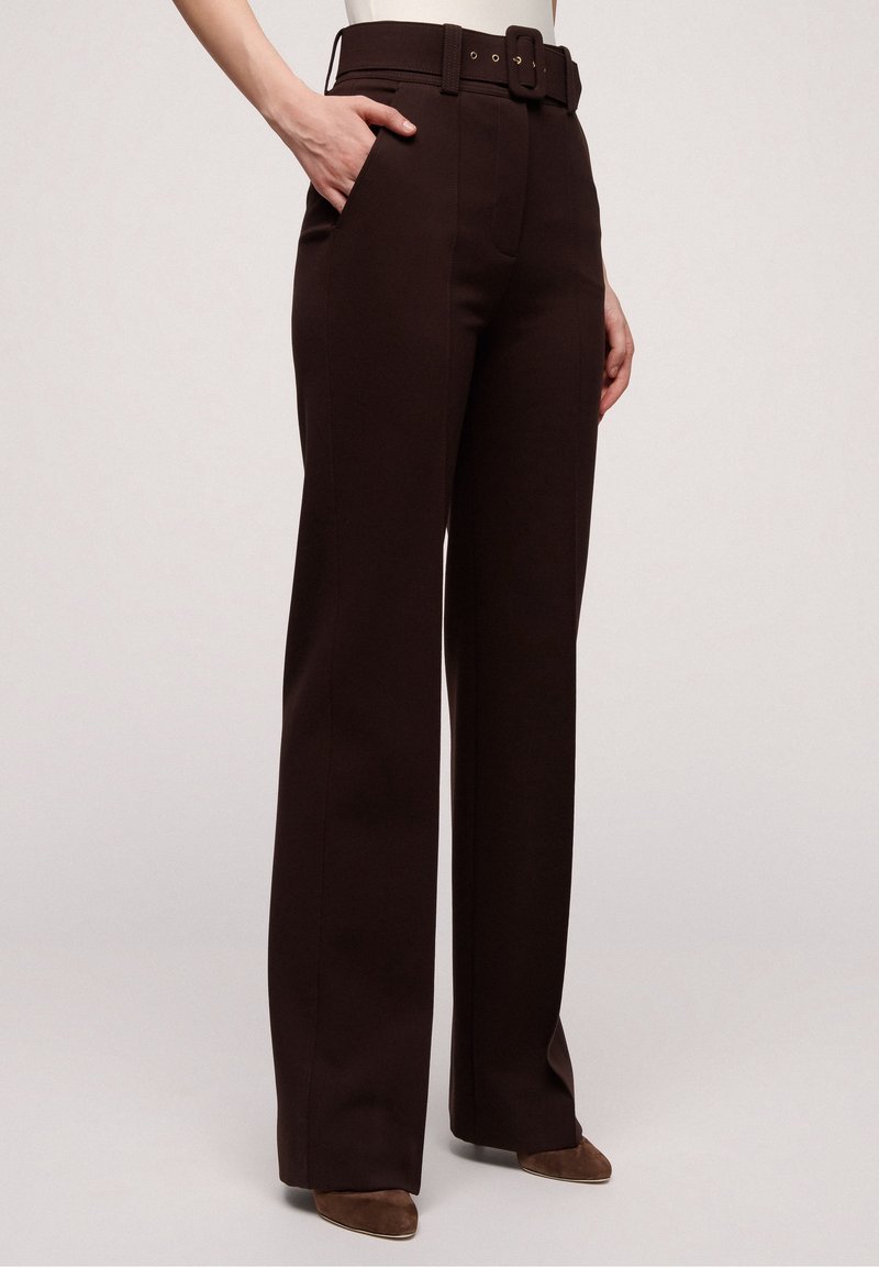 Femme portant un pantalon large taille haute marron foncé avec une ceinture assortie, main dans la poche, associée à des chaussures pointues marron.