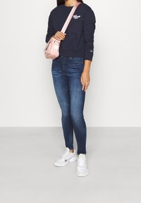 Marinblå sweatshirt, åtsittande blå jeans och vita sneakers. Rosa axelväska med en slät textur och justerbar rem. Enkel logotyp på t-shirten.