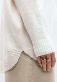 Šviesiai kremo spalvos megztinis su tekstūruotu paviršiumi, su dygsniuotomis rankogaliais ir šiek tiek išlenktu apačios kraštu, derinamas su smėlio spalvos audinio kelnėmis ir sluoksniuotais žiedais.