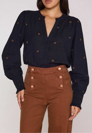 Kobieta ubrana w czarną bluzkę z długim rękawem z małą pomarańczową naszywką, połączoną z brązowymi, wysokim stanem spodniami z przodu zapinanymi na guziki.