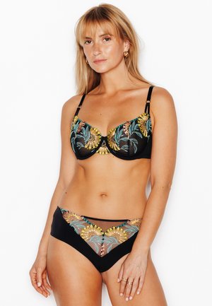 Femme debout, face à l’objectif, portant un ensemble soutiens-gorge et culotte noirs avec une broderie florale colorée, sur fond blanc uni.