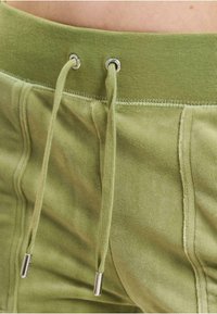 Zielone szorty z weluru z elastycznym paskiem, z metalowymi srebrnymi akcentami i miękką teksturą. Widoczne detale szwów podkreślają design.