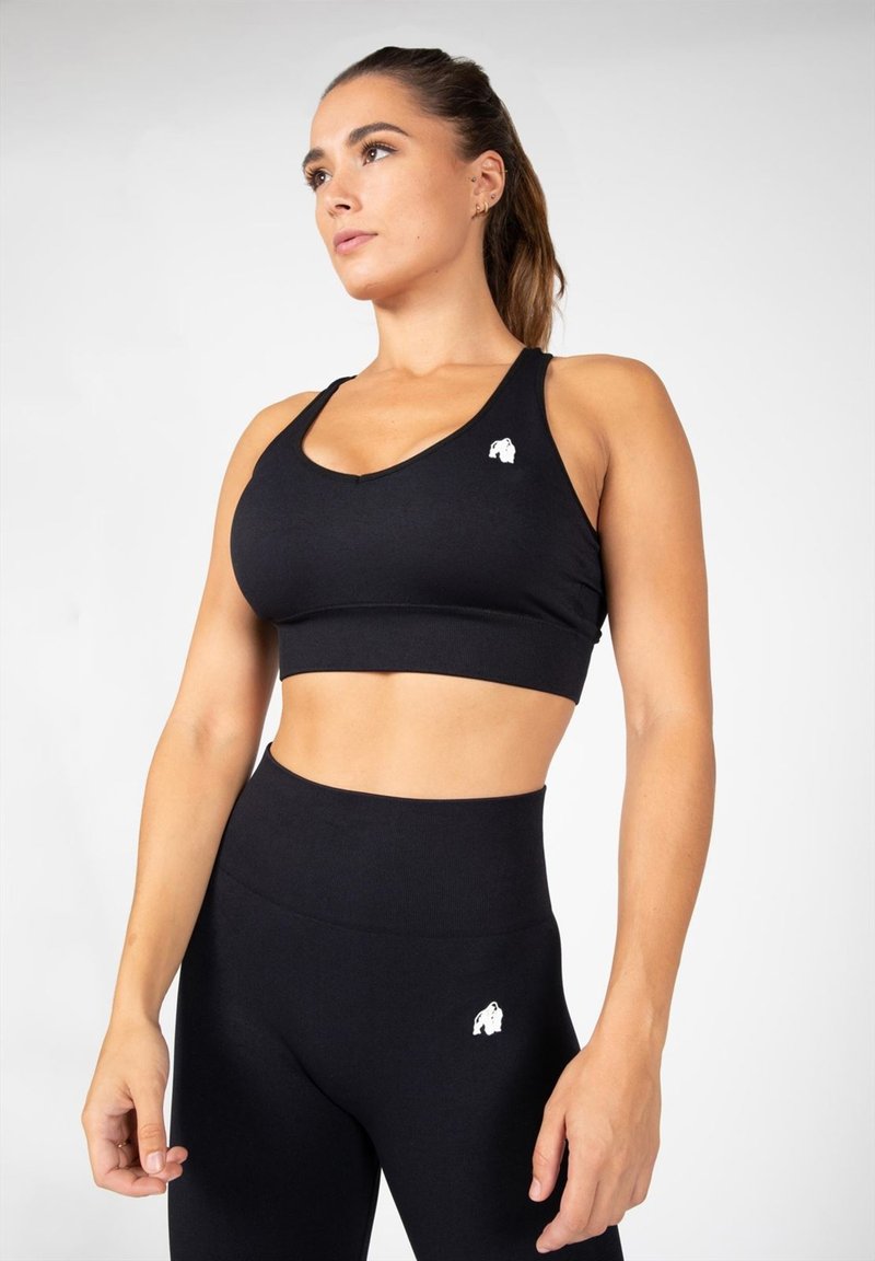 Zwarte sport-bh met een racerback-ontwerp, voorzien van een klein wit logo aan de linkerkant. De bijpassende hoge zwarte leggings tonen vergelijkbare branding.