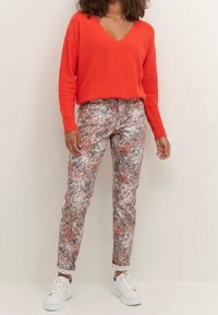 Kvinde iført en lys orange V-hals sweater, blomstermønstrede bukser og hvide sneakers, der står foran en lys baggrund.