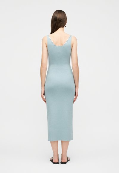 Γυναίκα που στέκεται πλάτη στην κάμερα, φορώντας αμάνικο γαλάζιο μίντι φόρεμα με ριμπ υφή και λεπτομέρειες με κουμπιά κατά μήκος του ντεκολτέ και μαύρα σανδάλια.