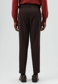 Bruine maatwerkbroek met rechte pijpen, voorzien van achterzakken en omgeslagen boorden, gecombineerd met donkere schoenen. Gladde stoftextuur.