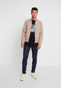 Veste bomber beige clair avec des poignets côtelés, portée sur un t-shirt graphique noir, accompagnée de jeans en denim foncé et de baskets blanches.