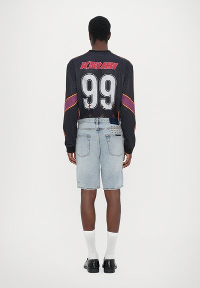 Cămașă neagră cu mânecă lungă, cu text roșu "K$UBI" și marele "99" alb, purtată cu pantaloni scurți din denim albastru deschis și pantofi negri. Sosete albe vizibile.