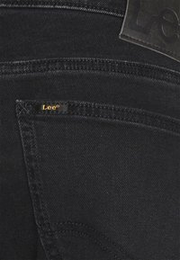 Gros plan sur la poche arrière d'un jean en denim noir présentant une petite étiquette de marque "Lee" et un patch en cuir sur la ceinture.