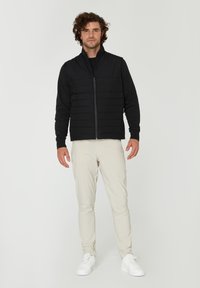 Svart vadderad väst med hög krage och zip-stängning, bärs över en svart skjorta. Ljust beige byxor och vita sneakers fulländar outfiten.