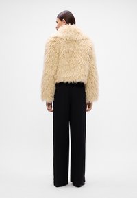 Faux fur jas in lichtbeige met een gestructureerd, shaggy oppervlak en een kraag. Gecombineerd met zwarte wijde broek voor contrast.