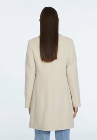 Mujer con cabello largo y lacio castaño, vestida con un abrigo de lana beige y jeans de color azul claro, vista desde atrás sobre un fondo blanco.