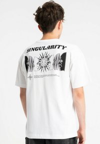 Młoda osoba z krótkimi włosami nosi białą koszulkę z napisem "SINGULARITY" oraz czarną liniową ilustracją twarzy wokół słońca na plecach.