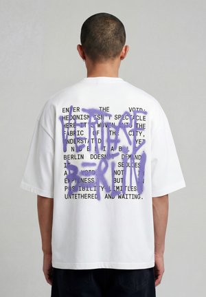 Persona vista da dietro che indossa una maglietta bianca oversize con testo nero e grande scritta in stile graffiti viola "VERBAL BARRI" sul retro.