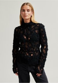 Černý texturovaný pletený svetr s dlouh�ými rukávy, který má vysoký límec a otevřené vzory, pod kterými se odhaluje kůže, doplněný tmavými džíny.