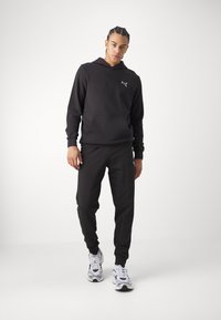 Sort hættetrøje og matchende joggers. Har en kængurulomme og et lille logo. Bæres med hvide og sorte sportsko.