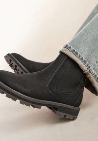 Schwarze Leder-Stiefeletten mit strukturiertem Finish, elastischen Seiteneinsätzen und robuster Gummisohle. Kombiniert mit hochgekrempelten hellblauen Jeans.