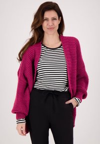 Fuchsia gebreide cardigan met geribbelde mouwen, gecombineerd met een zwart-wit gestreepte blouse en zwarte broek. Eenvoudig, losvallend ontwerp.