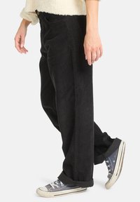 Pantalon en velours côtelé noir avec une coupe ample et des côtes verticales visibles, associé à des baskets en toile grises avec des semelles en caoutchouc blanches.