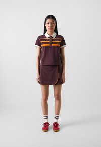 Barna póló ing narancssárga csíkokkal, fehér gallérral, barna szoknyával párosítva, amely narancssárga díszítéssel készült. Piros cipővel és fehér csíkos zoknival viselve.