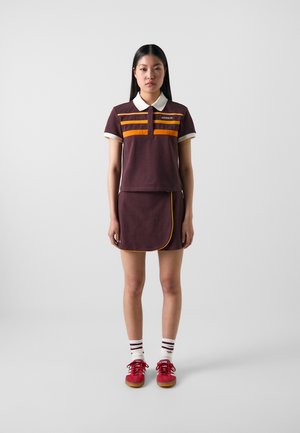 Polo shirt marron avec des rayures orange, col blanc, associé à une jupe marron avec passepoil orange. Porté avec des chaussures rouges et des chaussettes rayées blanches.