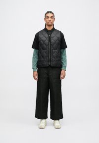 Chaleco acolchado negro sobre una camisa negra y una camiseta de manga larga color teal, combinado con pantalones anchos acolchados negros y zapatillas beige.