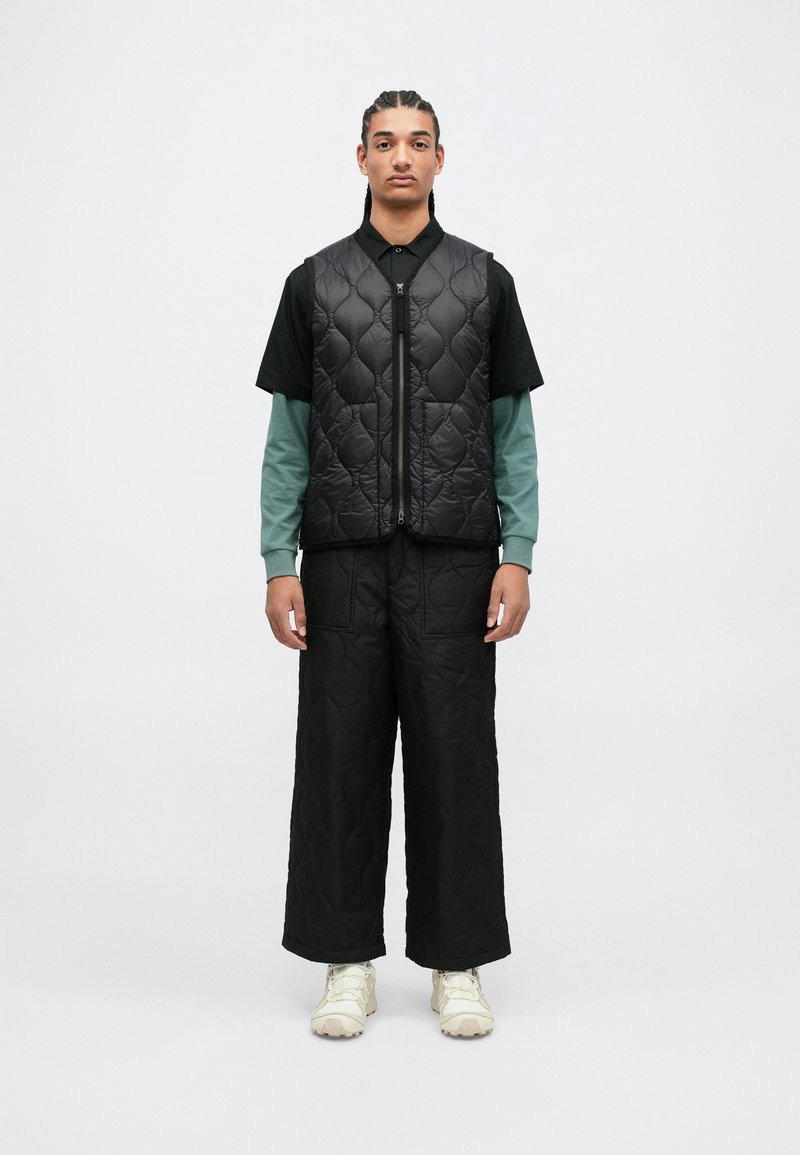 Chaleco acolchado negro sobre una camisa negra y una camiseta de manga larga color teal, combinado con pantalones anchos acolchados negros y zapatillas beige.