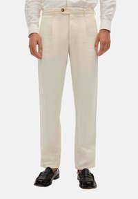 Beige Leinenhosen mit geradem Schnitt, ausgestattet mit zwei Vorderfalten, Gürtelschlaufen und einem einzelnen Knopfverschluss. Getragen mit schwarzen Slippern.