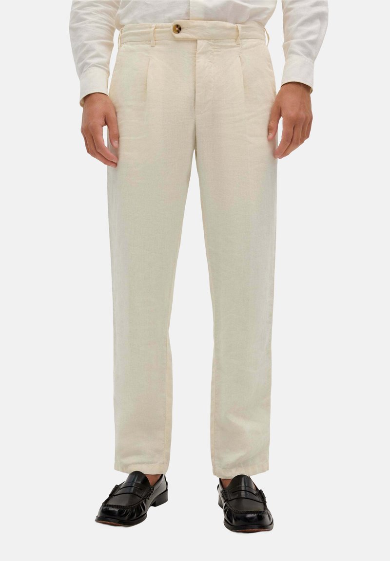 Beige Leinenhosen mit geradem Schnitt, ausgestattet mit zwei Vorderfalten, Gürtelschlaufen und einem einzelnen Knopfverschluss. Getragen mit schwarzen Slippern.
