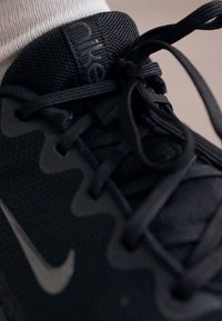 Scarpa sportiva nera con materiale in mesh, superficie testurizzata e lacci neri spessi con un logo sulla linguetta. Vista dall'alto.