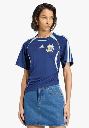 Donna che indossa una maglia da calcio Adidas Argentina blu con dettagli dorati e una gonna di jeans blu, in piedi con le mani rilassate ai lati.