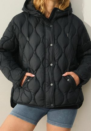 Doudoune noire matelassée avec fermeture à boutons et capuche, portée avec les mains dans les poches avant et associée à un short côtelé bleu.