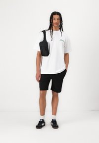 Bílá bavlněná tričko s zeleným textem, černé šortky, černá crossbody taška a černé tenisky s bílými pruhy a béžovými podrážkami.