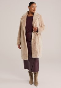 Manteau en fausse fourrure beige avec larges revers et fermeture par boutons à l'avant, associé à un haut bordeaux foncé et une jupe en cuir, terminé par des bottes beiges hautes jusqu'aux genoux.