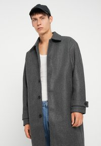 Grijze wollen jas met een knoopsluiting aan de voorkant, een grote kraag en opgerolde manchetten. Draagt over een witte tanktop, gecombineerd met blauwe jeans.